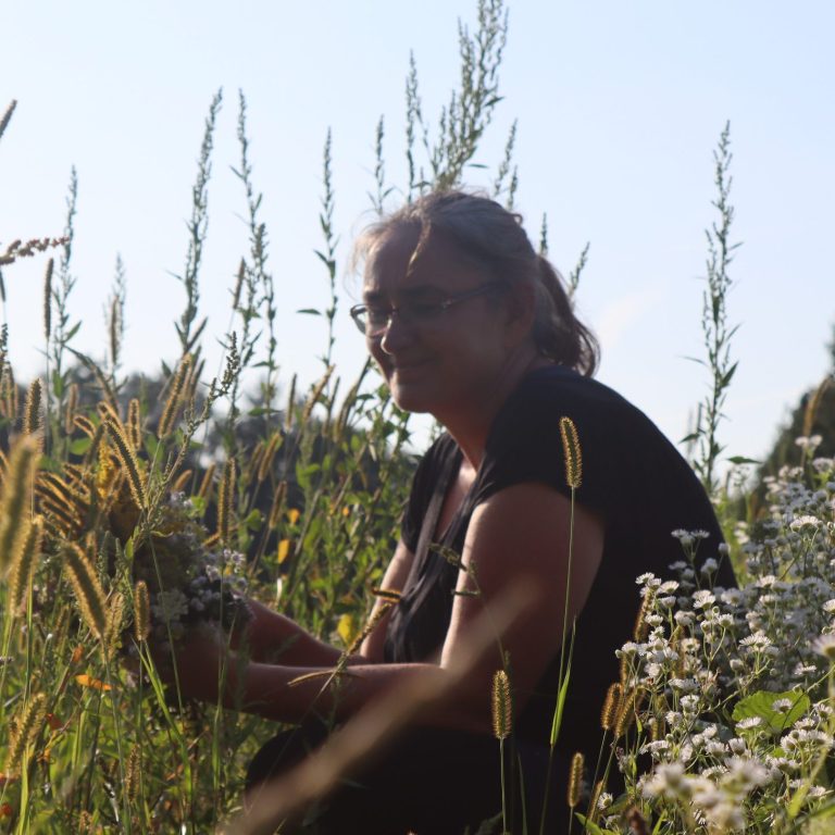 Frau mit Blumen im Feld, umgeben von hohen Gräsern und Blumen.