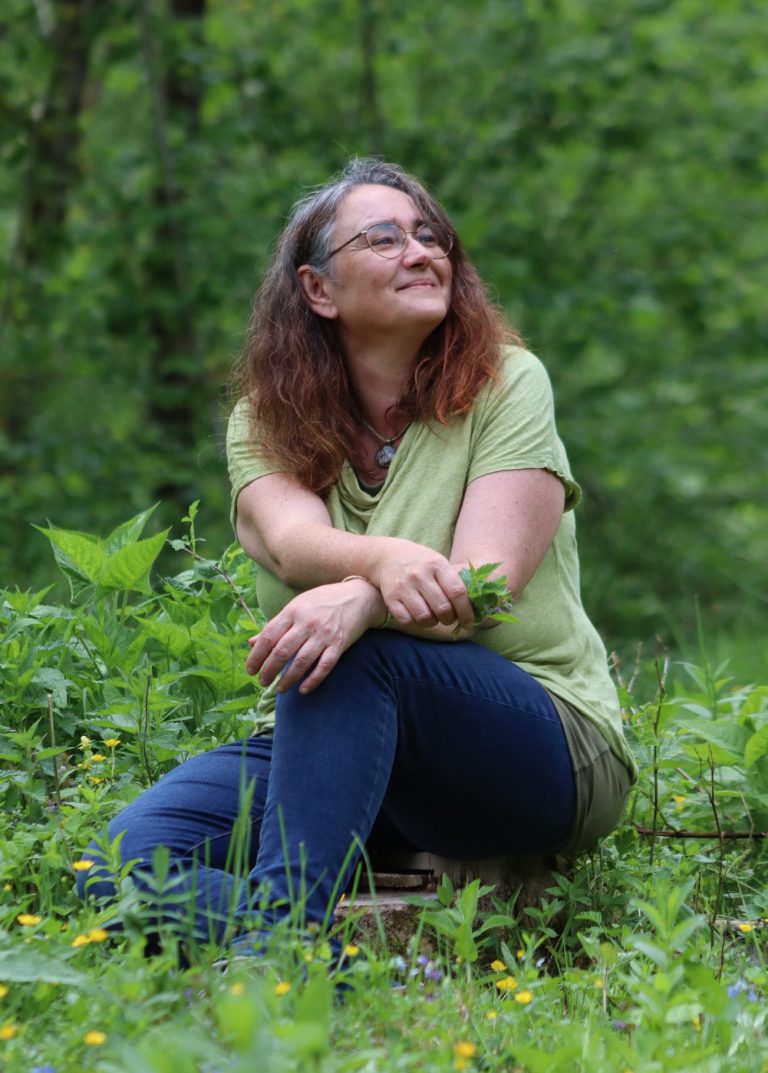 Frau mit langen, lockigen Haaren sitzt in einem grünen Wald und lächelt.