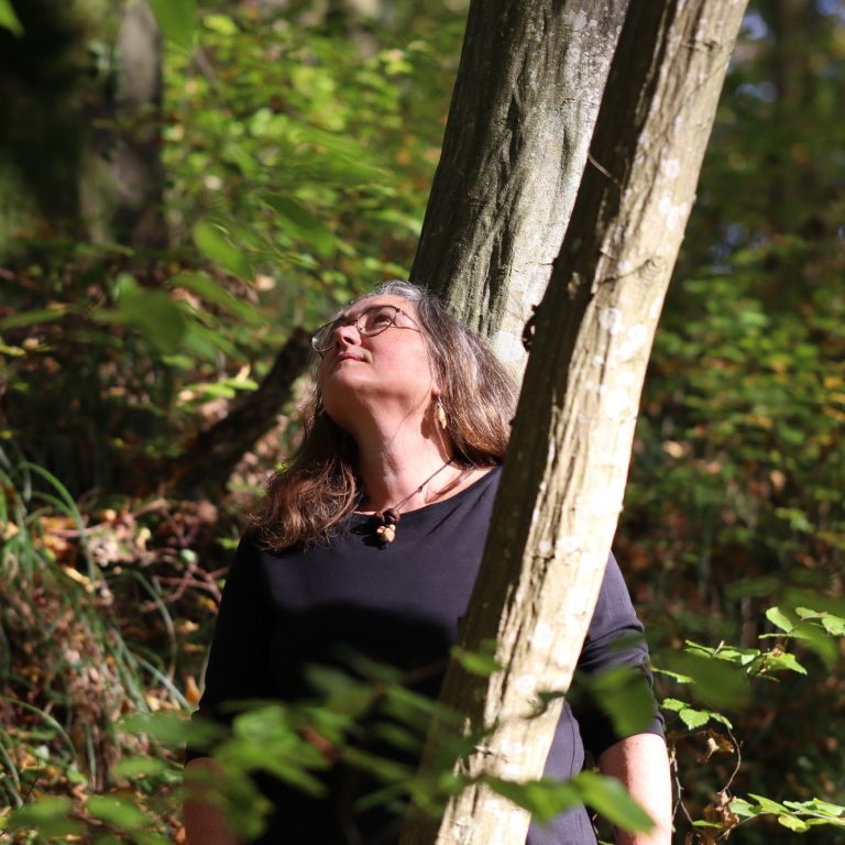 Eine Frau in einem dunklen Oberteil steht in einem Wald und schaut nach oben.